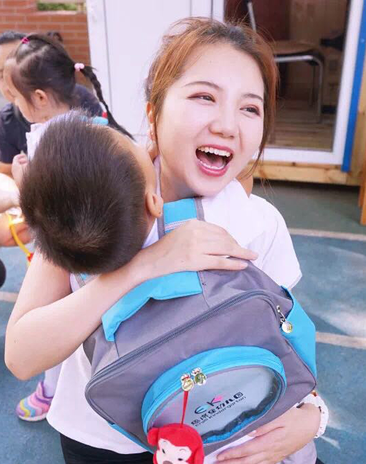 幼儿园教师拥抱背EK书包的孩子，温馨场景体现恩济里幼儿园校园文化建设