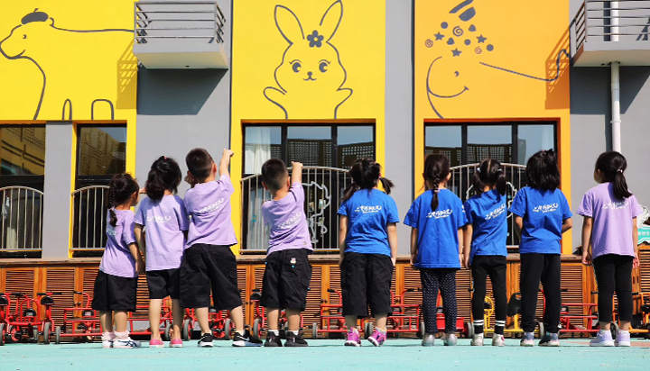 红英小学学生在黄色校园墙前排队，兔子小鹿墙绘环绕，阳光岛校园环境建设