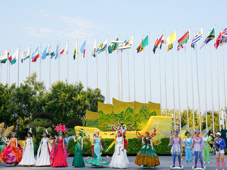 世园会国际区各国旗帜列阵，身着彩色民族服饰的人群在花卉景墙前合影