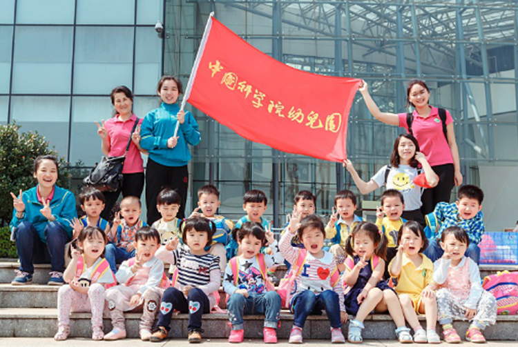 中科启元旗下中国科学院幼儿园师生在玻璃幕墙建筑前举红旗合影，展现教育集团品牌形象