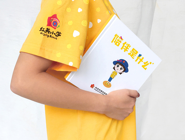 身穿红英小学黄色文化衫的学生臂夹'陪伴是什么'绘本，校园文化建设细节