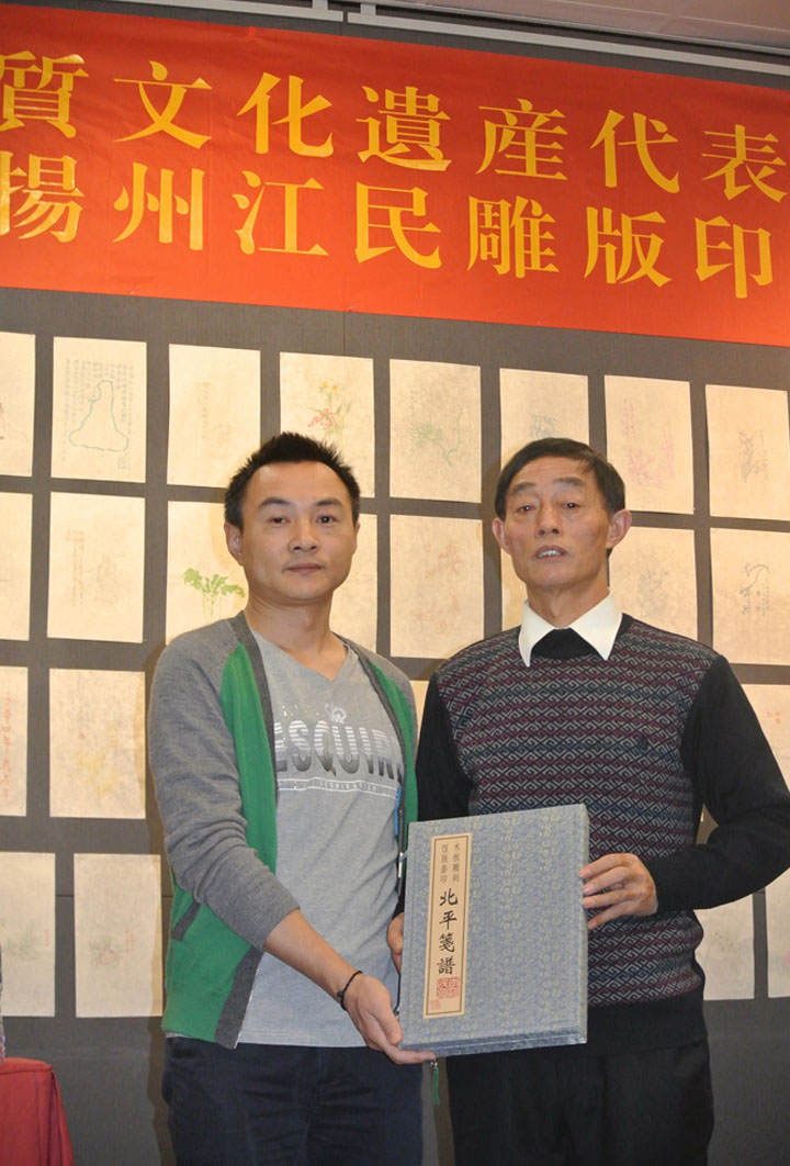 锦连环代表与扬州江民雕版印刷非遗大师合影，背景为非物质文化遗产展板