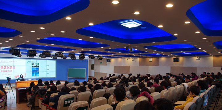 学校报告厅内锦连环校园文化培训现场侧拍，蓝色穹顶下师生满座聆听，前方大屏播放培训内容